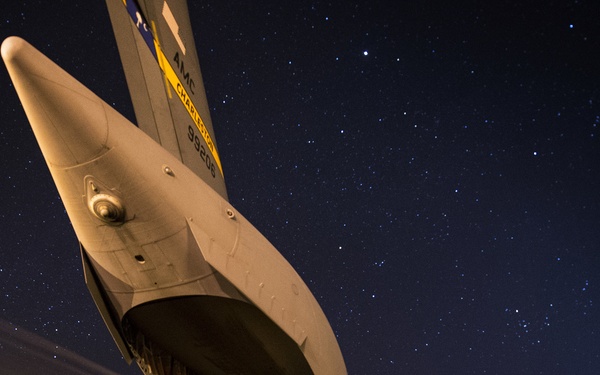 A large military cargo aircraft, identifiable as a C-17 Globemaster III based on its distinctive tail and markings, is photographed from a dramatic low-angle perspective at night on a flight line. The aircraft's rear cargo ramp is open and illuminated, with at least one ground crew member visible beneath the fuselage. The clear night sky filled with stars provides a striking backdrop to the operational scene.