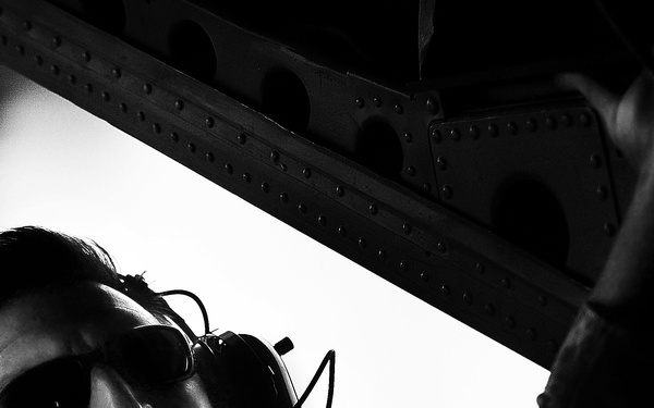 A black and white photograph taken from a low angle showing a person wearing aviation headset and sunglasses with their mouth open, appearing to be shouting or communicating. The dramatic upward angle reveals a riveted metal structural beam or aircraft component overhead. The high-contrast lighting creates a striking silhouette effect.