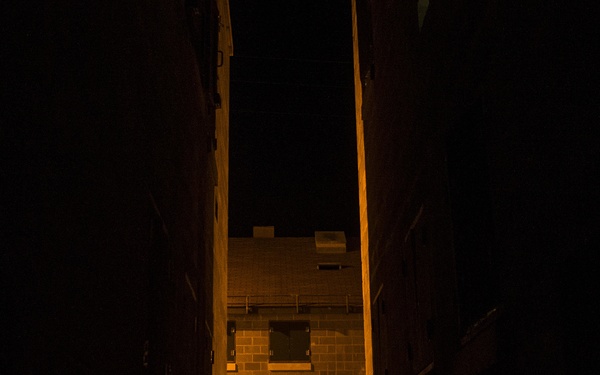 A nighttime image showing a silhouetted figure in tactical/military gear moving through a narrow passage or alleyway between structures. The scene is illuminated by an orange ambient light source, likely artificial lighting from the surrounding environment. The figure appears to be approaching a brick building with a door visible in the background.