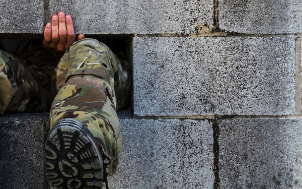 A person wearing military camouflage uniform and tactical boots is crawling through a rectangular opening in a concrete block wall. The image captures the lower body and legs of the individual mid-passage through the gap. This appears to be a military training obstacle course exercise.