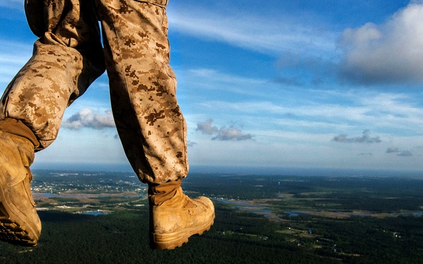 A military personnel wearing digital camouflage (MARPAT) uniform and tan combat boots is stepping off or standing at the edge of an aircraft ramp at high altitude. The perspective is a dramatic low-angle shot looking outward from the aircraft's open rear cargo ramp, revealing a vast aerial landscape below with forests, waterways, and scattered development. The image captures what appears to be a parachute jump or airborne operation in progress.