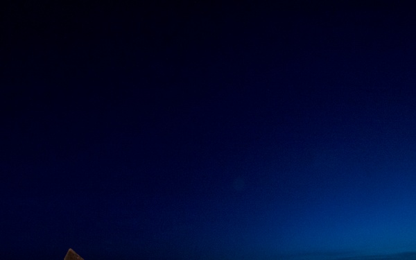 Two fishermen are working on a boat at night or during twilight, surrounded by a large catch of fish on the deck. They are wearing orange waterproof overalls and are positioned near stacked red fishing containers/barrels. The deep blue sky and calm sea horizon create a dramatic atmospheric backdrop.
