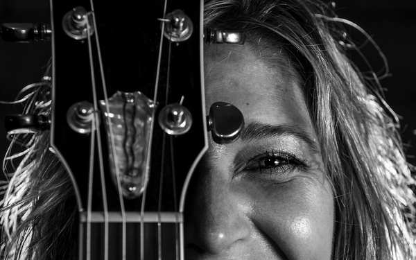 A black-and-white close-up portrait of a woman smiling broadly, with a guitar neck and headstock positioned vertically in front of her face, partially obscuring her left side. The guitar strings run across her face, creating a layered compositional effect. The image is a stylized artistic portrait combining the subject with her instrument.