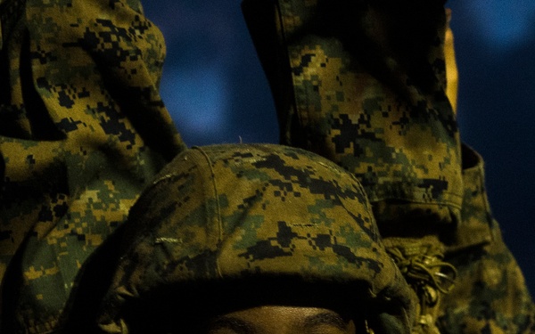 A close-up portrait of a person wearing MARPAT (Marine pattern) digital camouflage uniform and helmet, photographed at dusk or nighttime. The subject appears to be a military service member, likely a U.S. Marine, with sweat visible on their face suggesting physical exertion. The image is dramatically lit, highlighting the subject's face against a dark background with other camouflage-clad figures visible behind them.