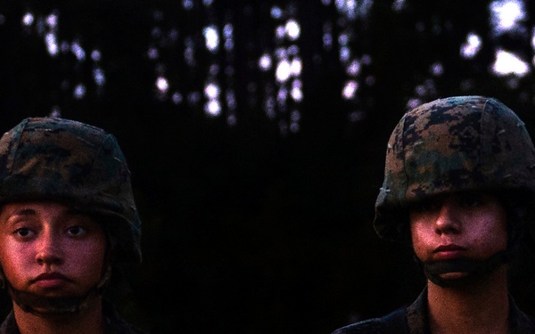 Four female military personnel are standing in formation outdoors during low-light conditions, wearing camouflage uniforms and combat helmets. They appear to be at attention or in a formal military stance, with a dark treeline visible in the background. The image appears to document a military training exercise or formation event.