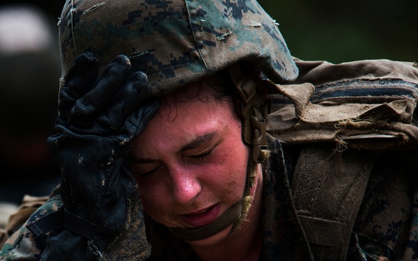 A military service member in US Marine Corps MARPAT digital camouflage uniform is photographed in a prone position during what appears to be a training exercise. The individual is visibly exhausted or distressed, covered in mud and dirt, holding a rifle while wearing full combat gear including helmet and pack. The close-up shot captures the physical and emotional strain of the training environment.