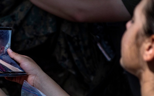 A person wearing military camouflage and an olive green t-shirt is holding and looking at a small collection of personal photographs. The top photo visible shows two people sharing a kiss. A clear plastic zip-lock bag containing additional photos or items is visible in their lap.