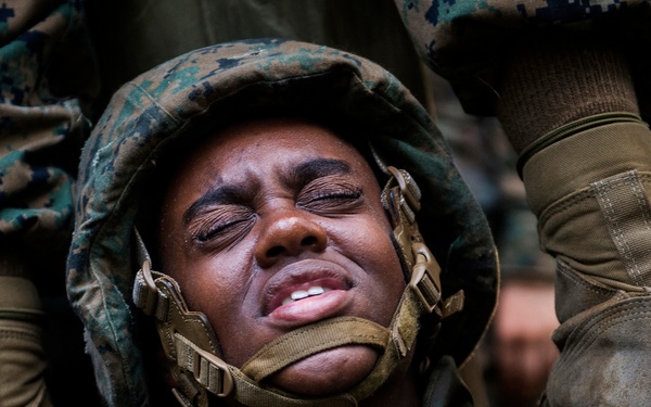 A close-up photograph of a military service member in camouflage uniform and helmet gear, appearing to be under significant physical strain or exertion. The individual's eyes are closed tightly and facial expression suggests intense effort or stress. Surrounding figures in similar military attire are partially visible, suggesting a group training or exercise scenario.