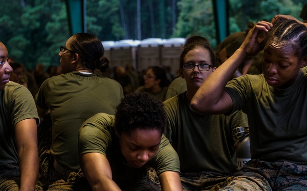 A group of female military recruits or trainees in olive drab t-shirts and camouflage trousers are resting during what appears to be a training exercise or field operation. One recruit is tending to another's foot, likely treating a blister or injury, while others rest and attend to personal needs. A canteen labeled 'MALONEY' is visible on the ground among the group.
