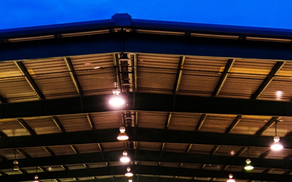 A large military formation is assembled under an open-sided covered pavilion or hangar structure at dusk or nighttime. Hundreds of personnel in camouflage uniforms are arranged in a horseshoe or U-shaped formation, with two individuals in lighter-colored uniforms standing at the center, one appearing to address the group. The scene is illuminated by overhead lights mounted to the metal roof structure.