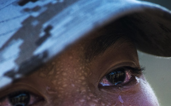 An extreme close-up photograph of a person wearing a digital camouflage military cap, with their face covered in water droplets and tears streaming down their cheeks. The eyes appear red and irritated, suggesting possible exposure to an irritant such as pepper spray or OC (oleoresin capsicum) spray, commonly used in military or law enforcement training. The image is emotionally intense and appears to document a training exercise or stress-inducing scenario.