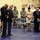 A military ceremony is taking place in what appears to be a gymnasium or armory, with U.S. Army personnel in dress uniforms and civilians standing with bowed heads in a moment of prayer or reflection. Three senior non-commissioned officers in Army Service Uniforms (blue dress) are prominently positioned in the foreground, while soldiers in ACU (Army Combat Uniform) and civilian attendees stand in the background. The setting and posture of participants suggest a formal military event such as a retirement, promotion, or memorial ceremony.