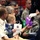 A military ceremony is taking place, likely at Raymond W. Bliss Army Health Center based on the visible program/pamphlet. Attendees seated in rows include uniformed military personnel and civilians, with a small toddler present. A person wearing white gloves is presenting gift bags to a seated elderly woman.