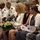 A formal military ceremony is taking place, with a mixed audience of uniformed military personnel and civilians seated in rows. Several women in the foreground hold yellow rose bouquets and printed programs, suggesting they are honored guests or family members. The setting appears to be an indoor venue such as a community center or military hall.