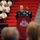 A U.S. Army soldier in dress blues is speaking at a podium draped with a burgundy cloth bearing the 'Service for Health' insignia, consistent with a U.S. Army Medical Command ceremony. The event appears to be a formal military ceremony held in a gymnasium or arena, with uniformed personnel seated on red bleachers in the background. An audience is visible in the foreground, watching the proceedings.