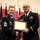 Two U.S. Army soldiers in dress blue uniforms are shaking hands while jointly holding a framed certificate or award document. The soldier on the left, identified by his name tape as 'NOETZELMAN,' holds the rank of Sergeant Major (indicated by his rank insignia). The ceremony appears to be a formal military awards presentation taking place in a hall decorated with multiple military unit flags.