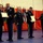 A formal U.S. Army military ceremony is taking place in an indoor venue, likely an armory or gymnasium. Five soldiers in dress uniforms are participating in what appears to be an awards or retirement ceremony, with framed certificates or citations being presented. State and unit flags are displayed in the background against a red and white backdrop.