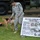 A U.S. Army Military Police soldier is handling a Military Working Dog (MWD) during what appears to be a public demonstration or display event. The soldier, wearing ACU (Army Combat Uniform) with an 'MP' patch, stands beside a Belgian Malinois-type dog near an informational sign about Military Working Dog Detection. The setting includes military vehicles in the background and educational materials about MWD capabilities.