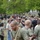 A group of U.S. Army personnel and civilians are participating in an outdoor team activity, likely a tug-of-war competition on a grassy field. The foreground shows a smiling soldier in a U.S. Army branded olive green t-shirt and red track pants gripping a thick rope. A large crowd of spectators and participants in military and civilian attire is visible in the background.