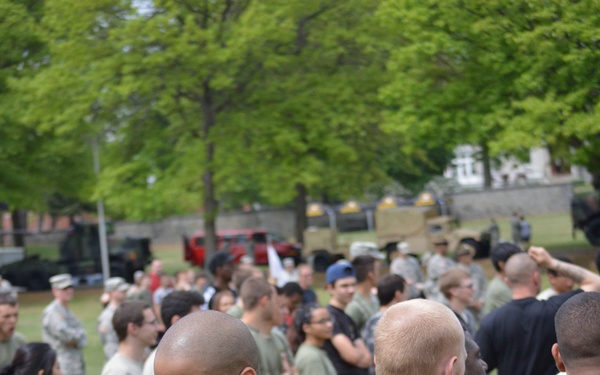 A group of U.S. Army personnel and civilians are participating in an outdoor team activity, likely a tug-of-war competition on a grassy field. The foreground shows a smiling soldier in a U.S. Army branded olive green t-shirt and red track pants gripping a thick rope. A large crowd of spectators and participants in military and civilian attire is visible in the background.