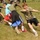 A group of young people are engaged in a tug-of-war competition on a grass field during what appears to be an outdoor school or community event. Participants are leaning back and pulling hard on a thick rope, showing visible physical effort and strain. A crowd of spectators and additional participants can be seen in the background.