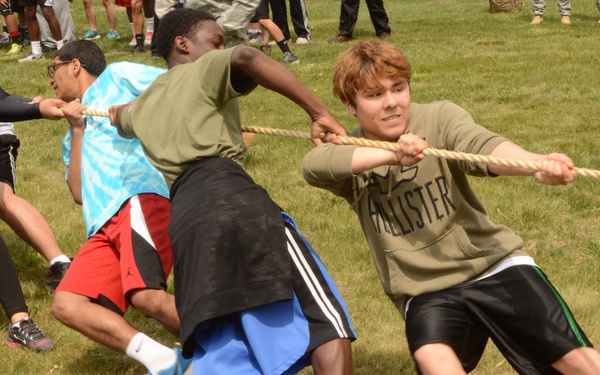 A group of young people are engaged in a tug-of-war competition on a grass field during what appears to be an outdoor school or community event. Participants are leaning back and pulling hard on a thick rope, showing visible physical effort and strain. A crowd of spectators and additional participants can be seen in the background.
