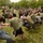 A large group of people, including civilians and military personnel in Army combat uniforms, are participating in a tug-of-war competition on a grassy field. The event appears to be a military community or recruitment event, with military vehicles including helicopters and HMMWVs visible in the background. A uniformed soldier observes from the right side while spectators watch the competition.
