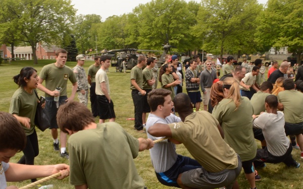 A large group of people, including civilians and military personnel in Army combat uniforms, are participating in a tug-of-war competition on a grassy field. The event appears to be a military community or recruitment event, with military vehicles including helicopters and HMMWVs visible in the background. A uniformed soldier observes from the right side while spectators watch the competition.