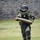 A person wearing a full EOD (Explosive Ordnance Disposal) bomb suit is carrying a large cylindrical olive-drab object across a grass field. The individual is dressed in a complete blast-protective ensemble including helmet with visor, padded torso protection, and reinforced leg guards. The setting appears to be a training exercise or demonstration near a stone wall structure.