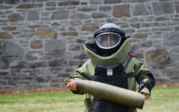 A person wearing a full EOD (Explosive Ordnance Disposal) bomb suit is carrying a large cylindrical olive-drab object across a grass field. The individual is dressed in a complete blast-protective ensemble including helmet with visor, padded torso protection, and reinforced leg guards. The setting appears to be a training exercise or demonstration near a stone wall structure.