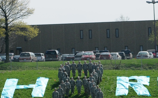 A group of military personnel in camouflage uniforms are standing together on a green grass field, positioned between large light-blue letters spelling 'SHARP'. The letters appear to be made from fabric or tarps laid on the ground, forming a display likely related to the U.S. Army's SHARP (Sexual Harassment/Assault Response and Prevention) program. The scene appears to be a formal awareness or promotional event photographed from a distance.
