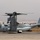A U.S. Marines MV-22 Osprey tiltrotor aircraft is parked on a tarmac, identifiable by its distinctive twin-rotor configuration with rotors folded in helicopter mode. The aircraft bears markings for Marine Medium Tiltrotor Squadron VMM-266, with bureau number 8303 and modex '00'. The background shows a hazy urban environment with low-rise buildings and arid terrain, suggesting a location in the Middle East or North Africa.