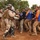 The image depicts what appears to be a military training exercise involving riot control or crowd management scenarios. Soldiers in camouflage uniforms with riot gear, shields, and batons are engaging with a group of civilians wearing blue and orange jackets, some labeled 'GER'. The setting is an outdoor dirt area with trees in the background, consistent with a training environment.
