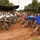 The image depicts a military riot control training exercise taking place on a dirt field. A line of soldiers wearing white helmets and carrying riot shields advances against a group of role-players simulating a crowd, while a larger audience of uniformed military personnel observes from a covered grandstand in the background. The exercise appears to be a joint or multinational training demonstration.