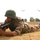 A soldier in camouflage uniform and green steel helmet is lying prone on dry grassland in a firing position, holding what appears to be an AK-pattern rifle. In the background, two tiltrotor aircraft — identifiable as V-22 Osprey type — are visible in flight. The scene appears to depict a military exercise or training operation.