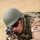 A soldier in prone firing position aims a rifle with an optical scope directly toward the camera. The soldier wears a Dutch military flag patch on the right sleeve, indicating affiliation with the Royal Netherlands Army. The image captures a training or operational scenario with the soldier in full combat gear.