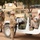 The image depicts what appears to be a military training exercise or checkpoint scenario involving multiple soldiers. Personnel in different uniforms are interacting around military vehicles, including a white Humvee and a tan/desert-colored armored vehicle. Several individuals in the white vehicle have their hands raised, suggesting a simulated detainment or vehicle stop drill.