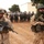 Two armed military personnel stand in the foreground in a desert environment, facing the camera while holding assault rifles. Behind them, a large formation of personnel carrying riot shields is visible, suggesting a training exercise or crowd control operation. The setting appears to be a forward operating base or training facility in an arid region.