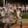 Military personnel in full riot control gear, including helmets with face shields, body armor, knee pads, and transparent shields, are conducting a demonstration or training exercise. A formation of soldiers holds riot shields in a line while one individual moves forward carrying a baton. An audience of uniformed military observers, including personnel in various camouflage patterns and at least one in green dress uniform, watches from bleachers in the background.