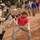 The image depicts what appears to be a military training exercise involving riot control or crowd management scenarios. A soldier in full riot gear including a face shield and baton faces off against a role-player in civilian-style clothing in a combative stance. Multiple military and civilian personnel observe or participate in similar drills in the background on a dirt training ground.