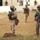 The image shows multiple military personnel in a field or compound area, wearing different camouflage uniforms and carrying rifles. At least two distinct uniform patterns are visible — MARPAT (Marine pattern) and UCP (Army digital camouflage) — suggesting a joint or multinational military exercise or operation. Additional soldiers in what appear to be foreign military uniforms are visible in the background.