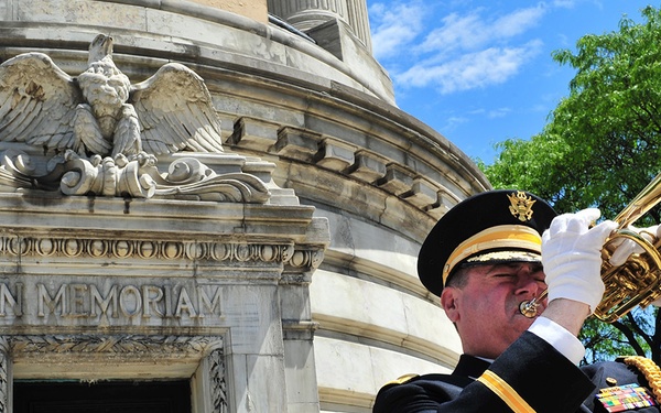 A U.S. Army soldier in dress uniform plays a bugle in front of a stone memorial monument inscribed with 'IN MEMORIAM.' The scene appears to be a military memorial ceremony, likely on Memorial Day or a similar commemorative occasion. Floral wreaths in red, white, and blue are visible at the base of the monument.