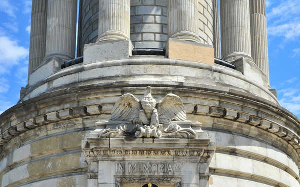 A military officer in dress uniform stands at attention on the steps of a neoclassical memorial monument, holding a bugle. The monument bears the inscription 'IN MEMORIAM' above its entrance, flanked by an eagle sculpture and decorative wreaths in red, white, and blue. The setting strongly suggests a memorial ceremony, likely Veterans Day or Memorial Day.