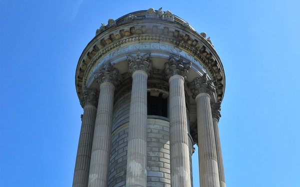 Two men in dark suits ascend stone steps toward a large circular neoclassical memorial monument, carrying or accompanying a large floral wreath with red, white, and blue ribbons. A third figure in a white naval or military uniform is visible near the monument's entrance. The scene appears to be a formal wreath-laying ceremony at what appears to be the Soldiers' and Sailors' Monument in New York City.