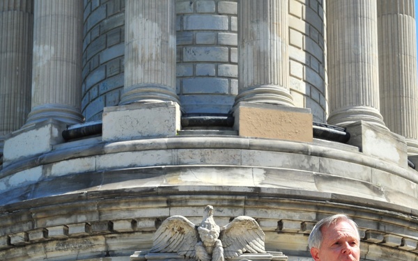 A man in a dark suit with a blue tie is speaking at a wooden podium with microphones in front of a large neoclassical stone monument. The monument features classical columns, an eagle relief sculpture, and the inscription 'IN MEMORIAM', suggesting a memorial or commemorative ceremony. The setting is outdoors under a clear blue sky.