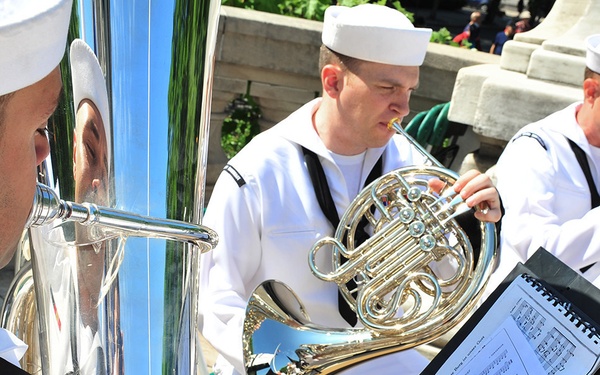 Three U.S. Navy musicians from 'Navy Band Northeast' are performing outdoors in white dress uniforms and white sailor caps. The musicians are playing brass instruments with sheet music on a stand visible in the foreground. The performance appears to be taking place at a public outdoor venue, likely a memorial or civic space.