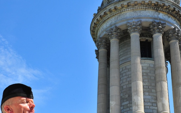 A bagpiper dressed in traditional Scottish attire performs outdoors in front of a large neoclassical stone monument or memorial tower. The structure features Corinthian columns and an inscription reading 'TO THE MEMORY OF' along its frieze. The scene appears to be a formal commemorative or ceremonial event on a clear, sunny day.