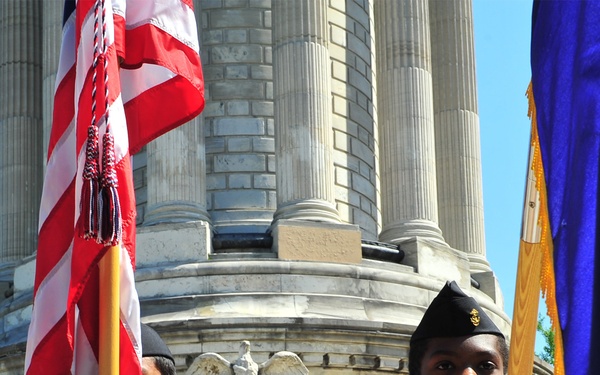 A color guard ceremony is taking place at what appears to be a war memorial monument, with young cadets in tan uniforms holding flags including the American flag. A Navy sailor in dress whites is speaking at a podium in the background. The stone monument behind them features an eagle relief sculpture and the partial inscription 'IN MEMORIA'.