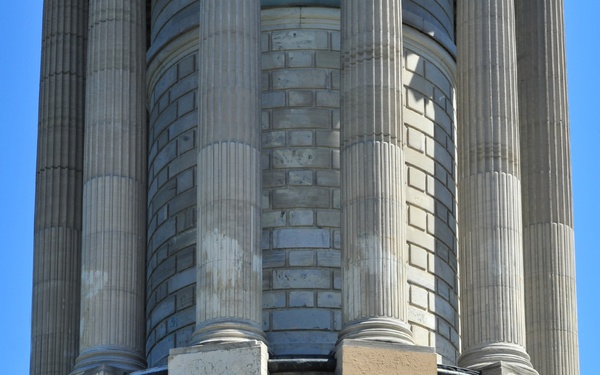 A man in a dark suit is speaking at a wooden podium equipped with multiple microphones, positioned in front of a large neoclassical stone monument or memorial structure. The building features fluted columns, ornate stonework, and an eagle relief carving visible behind the speaker. The setting suggests a formal public ceremony or commemorative event held outdoors under clear blue skies.