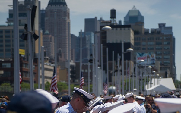 A military ceremony is taking place outdoors in an urban setting, with numerous service members in white Navy dress uniforms and covers folding or handling a large American flag. The event appears to be a formal flag-folding ceremony, likely associated with Fleet Week or a similar military commemoration given the New York City skyline visible in the background. Multiple branches of service appear to be represented, including Navy and Coast Guard personnel.